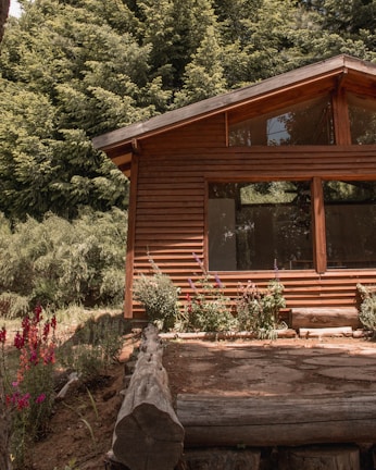 A wooden cabin with large windows is surrounded by lush green trees and colorful flowers. The wooden structure blends naturally with the forest scenery, adding to a rustic and peaceful environment.