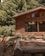 A wooden cabin with large windows is surrounded by lush green trees and colorful flowers. The wooden structure blends naturally with the forest scenery, adding to a rustic and peaceful environment.
