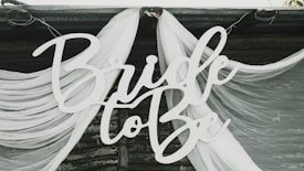 A decorative sign with the text 'Bride to Be' is displayed against a backdrop of flowing white fabric draped elegantly. The fabric appears sheer and is arranged in soft, graceful folds.