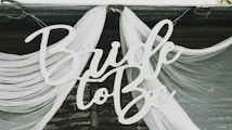 A decorative sign with the text 'Bride to Be' is displayed against a backdrop of flowing white fabric draped elegantly. The fabric appears sheer and is arranged in soft, graceful folds.
