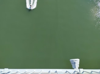 A small watercraft is moving across a wide expanse of green water, leaving a white wake behind. The viewpoint is elevated, looking down on the vessel. In the bottom part of the image, a structure with a metal railing and a spotlight is visible.
