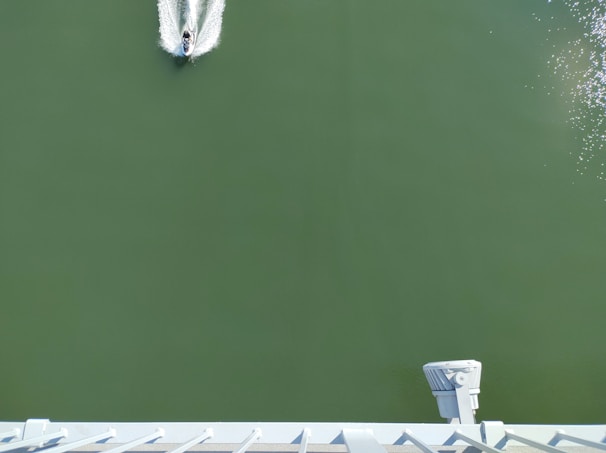 A small watercraft is moving across a wide expanse of green water, leaving a white wake behind. The viewpoint is elevated, looking down on the vessel. In the bottom part of the image, a structure with a metal railing and a spotlight is visible.