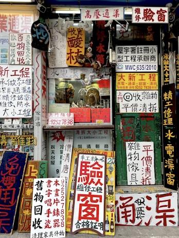 A densely packed storefront with a variety of colorful signs in different sizes and fonts, all with Chinese characters. The signs are stacked and layered, some leaning against others, suggesting a busy, eclectic business. The storefront is partially obscured by the abundance of signage, with a few visible objects like water pipes and a reflective grill.
