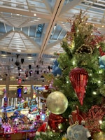A festive Christmas tree adorned with colorful ornaments, including a large red icicle, a silver globe, and a variety of playful figures like a teddy bear and a blue character. The background features decorative lighting and a lively shopping mall environment with holiday-themed displays.