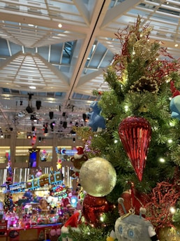A festive Christmas tree adorned with colorful ornaments, including a large red icicle, a silver globe, and a variety of playful figures like a teddy bear and a blue character. The background features decorative lighting and a lively shopping mall environment with holiday-themed displays.