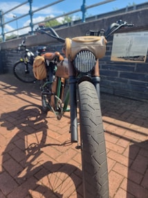 A vintage-style bicycle is parked on a brick pathway near a stone wall. The bike has leather saddlebags and a headlight resembling a motorcycle, with a front view focusing on its large tire. Another bicycle is visible in the background, along with a historical informational plaque mounted on the wall.