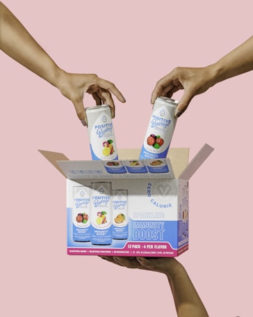 Two hands are placing cans of Positive Beverage into an open cardboard box against a soft pink background. The box features various fruit illustrations and prominently advertises the drinks as zero calorie and sparkling with an immunity boost. A label at the bottom indicates it is a 12 pack with four flavors.