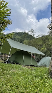 Several green tents are set up in a grassy field, surrounded by lush greenery and hills. The sky is partially cloudy, creating a serene and peaceful outdoor camping scene.