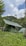 Several green tents are set up in a grassy field, surrounded by lush greenery and hills. The sky is partially cloudy, creating a serene and peaceful outdoor camping scene.