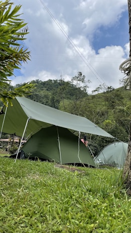 Several green tents are set up in a grassy field, surrounded by lush greenery and hills. The sky is partially cloudy, creating a serene and peaceful outdoor camping scene.