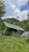 Several green tents are set up in a grassy field, surrounded by lush greenery and hills. The sky is partially cloudy, creating a serene and peaceful outdoor camping scene.