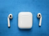 A pair of wireless earphones are placed on each side of a square-shaped charging case, all of which are white. These objects are set against a bright blue surface.