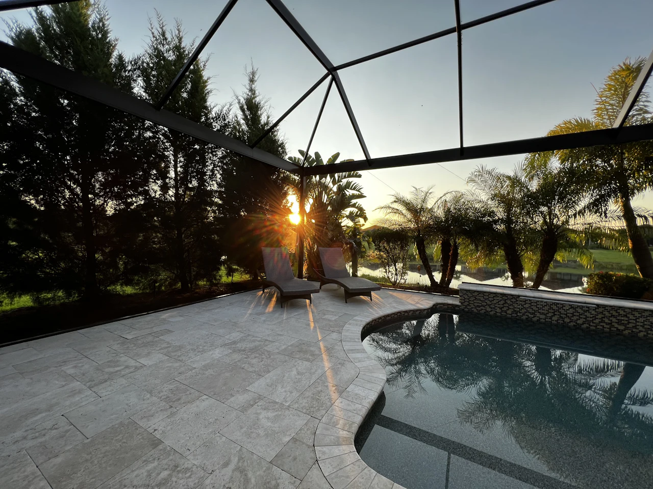A screened pool lanai at sunset with lounge chairs and a travertine deck