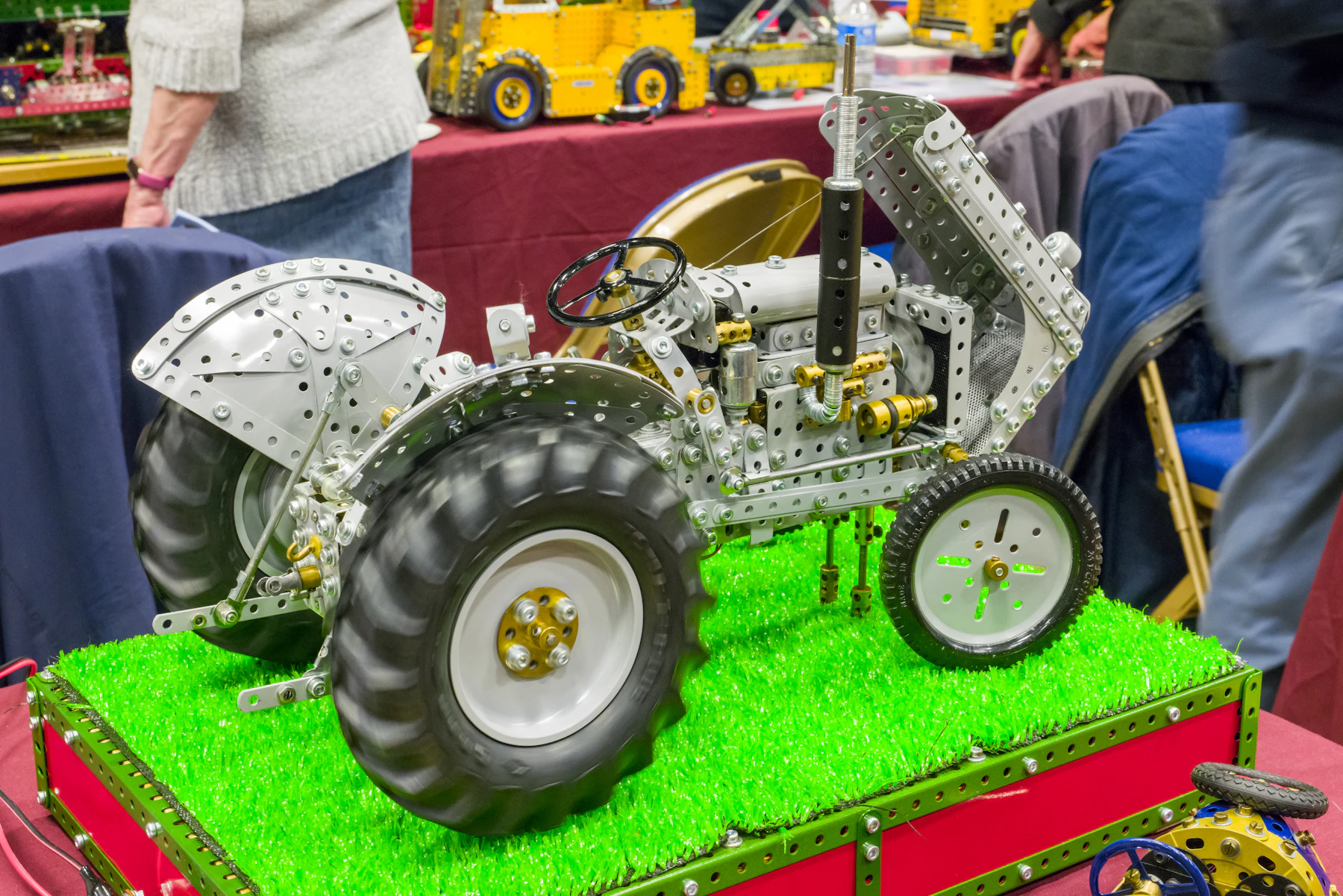 A large gray tractor built from perforated metal plates and brass fittings on a green display base at an exhibition