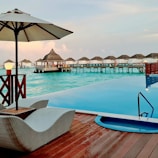 An infinity pool overlooks a tranquil ocean with overwater bungalows in the distance. A cozy lounge chair and parasol are positioned by the pool, set on a wooden deck. The scene is illuminated by soft lighting, likely captured during twilight.