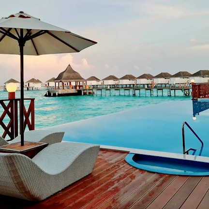 An infinity pool overlooks a tranquil ocean with overwater bungalows in the distance. A cozy lounge chair and parasol are positioned by the pool, set on a wooden deck. The scene is illuminated by soft lighting, likely captured during twilight.