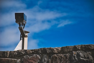 A surveillance camera mounted on a metal pole is positioned above a stone wall, with a clear blue sky in the background.