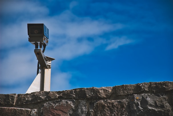 A surveillance camera mounted on a metal pole is positioned above a stone wall, with a clear blue sky in the background.