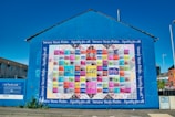 A large, vibrant mural painted on the side of a building displays a colorful patchwork quilt with various words and phrases like 'love,' 'hope,' and 'mother.' The mural is encased in a bright blue border with the text 'Women's Voices Matter... Equality for all.' The artwork is accompanied by informational text about 'The Women's Quilt' on an adjacent wall. Surrounding the mural are buildings, a clear sky, and a street with some greenery.
