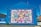 A large, vibrant mural painted on the side of a building displays a colorful patchwork quilt with various words and phrases like 'love,' 'hope,' and 'mother.' The mural is encased in a bright blue border with the text 'Women's Voices Matter... Equality for all.' The artwork is accompanied by informational text about 'The Women's Quilt' on an adjacent wall. Surrounding the mural are buildings, a clear sky, and a street with some greenery.