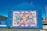 A large, vibrant mural painted on the side of a building displays a colorful patchwork quilt with various words and phrases like 'love,' 'hope,' and 'mother.' The mural is encased in a bright blue border with the text 'Women's Voices Matter... Equality for all.' The artwork is accompanied by informational text about 'The Women's Quilt' on an adjacent wall. Surrounding the mural are buildings, a clear sky, and a street with some greenery.