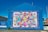 A large, vibrant mural painted on the side of a building displays a colorful patchwork quilt with various words and phrases like 'love,' 'hope,' and 'mother.' The mural is encased in a bright blue border with the text 'Women's Voices Matter... Equality for all.' The artwork is accompanied by informational text about 'The Women's Quilt' on an adjacent wall. Surrounding the mural are buildings, a clear sky, and a street with some greenery.