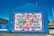 A large, vibrant mural painted on the side of a building displays a colorful patchwork quilt with various words and phrases like 'love,' 'hope,' and 'mother.' The mural is encased in a bright blue border with the text 'Women's Voices Matter... Equality for all.' The artwork is accompanied by informational text about 'The Women's Quilt' on an adjacent wall. Surrounding the mural are buildings, a clear sky, and a street with some greenery.