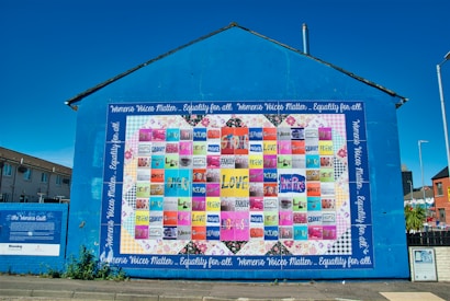 A large, vibrant mural painted on the side of a building displays a colorful patchwork quilt with various words and phrases like 'love,' 'hope,' and 'mother.' The mural is encased in a bright blue border with the text 'Women's Voices Matter... Equality for all.' The artwork is accompanied by informational text about 'The Women's Quilt' on an adjacent wall. Surrounding the mural are buildings, a clear sky, and a street with some greenery.