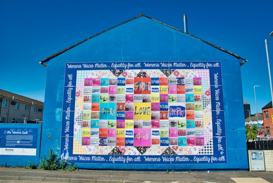 A large, vibrant mural painted on the side of a building displays a colorful patchwork quilt with various words and phrases like 'love,' 'hope,' and 'mother.' The mural is encased in a bright blue border with the text 'Women's Voices Matter... Equality for all.' The artwork is accompanied by informational text about 'The Women's Quilt' on an adjacent wall. Surrounding the mural are buildings, a clear sky, and a street with some greenery.