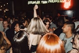 A crowded dance floor with people enjoying themselves, some raising their hands and others holding drinks. The atmosphere is lively, with a neon sign in the background reading 'Stranger Than Paradise.' The diversity of expressions and interactions among the crowd suggests a vibrant party scene.