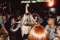 A crowded dance floor with people enjoying themselves, some raising their hands and others holding drinks. The atmosphere is lively, with a neon sign in the background reading 'Stranger Than Paradise.' The diversity of expressions and interactions among the crowd suggests a vibrant party scene.
