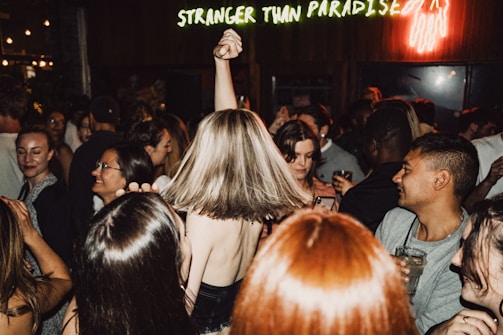 A crowded dance floor with people enjoying themselves, some raising their hands and others holding drinks. The atmosphere is lively, with a neon sign in the background reading 'Stranger Than Paradise.' The diversity of expressions and interactions among the crowd suggests a vibrant party scene.