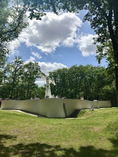 A modern sculpture made of what appears to be smooth, white material contrasts with the surrounding lush greenery. The artwork stands atop a winding structure with people sitting along its curve. The sky above is partly cloudy with patches of blue visible.