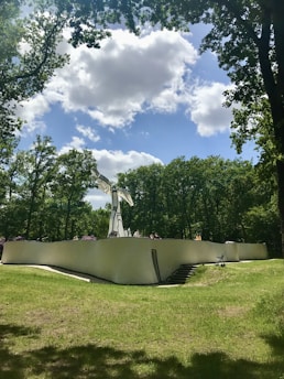 A modern sculpture made of what appears to be smooth, white material contrasts with the surrounding lush greenery. The artwork stands atop a winding structure with people sitting along its curve. The sky above is partly cloudy with patches of blue visible.