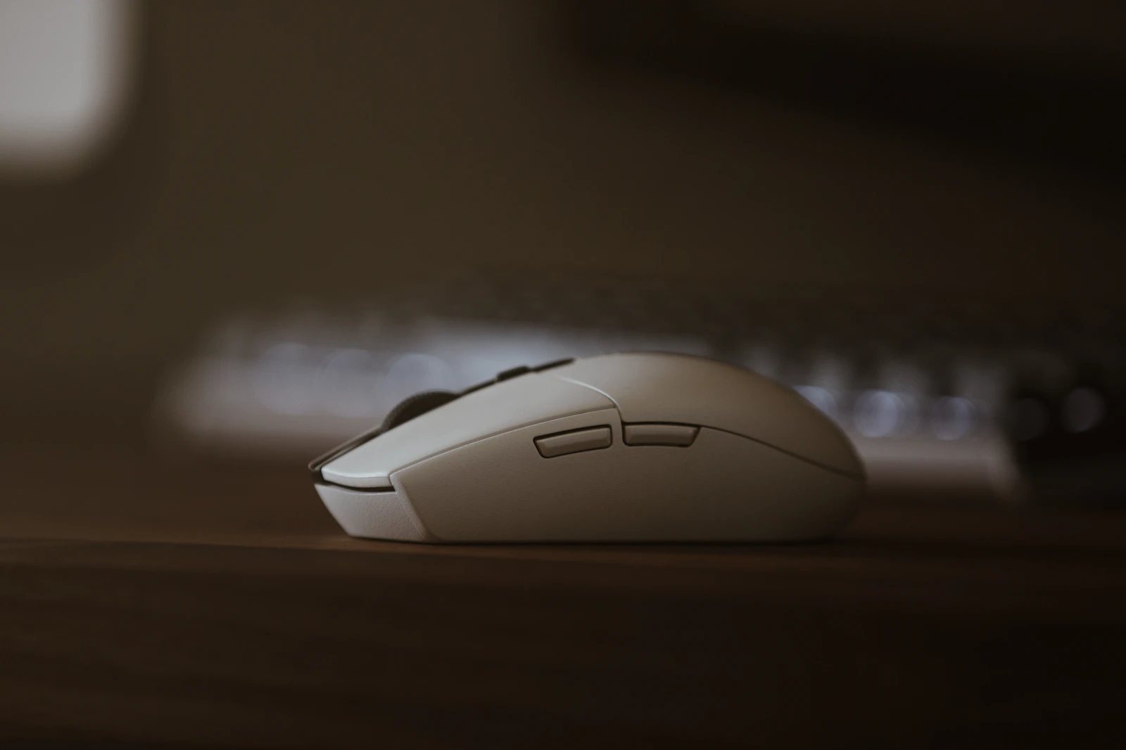 Close-up side profile of a compact white wireless computer mouse resting on a wooden desk with a keyboard blurred in the background