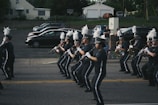 A marching band in uniform is performing on a street. The band members are holding musical instruments, such as clarinets and saxophones, and are in synchronized formation. They wear dark uniforms with white plumes on their hats. Parked cars and a small audience can be seen in the background.