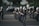 A marching band in uniform is performing on a street. The band members are holding musical instruments, such as clarinets and saxophones, and are in synchronized formation. They wear dark uniforms with white plumes on their hats. Parked cars and a small audience can be seen in the background.