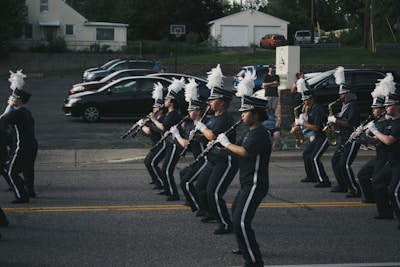 A marching band in uniform is performing on a street. The band members are holding musical instruments, such as clarinets and saxophones, and are in synchronized formation. They wear dark uniforms with white plumes on their hats. Parked cars and a small audience can be seen in the background.