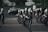 A marching band in uniform is performing on a street. The band members are holding musical instruments, such as clarinets and saxophones, and are in synchronized formation. They wear dark uniforms with white plumes on their hats. Parked cars and a small audience can be seen in the background.