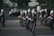 A marching band in uniform is performing on a street. The band members are holding musical instruments, such as clarinets and saxophones, and are in synchronized formation. They wear dark uniforms with white plumes on their hats. Parked cars and a small audience can be seen in the background.