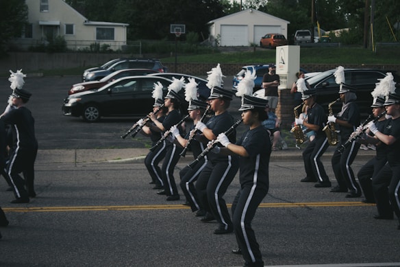 A marching band in uniform is performing on a street. The band members are holding musical instruments, such as clarinets and saxophones, and are in synchronized formation. They wear dark uniforms with white plumes on their hats. Parked cars and a small audience can be seen in the background.