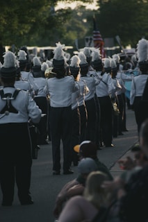 A marching band dressed in uniforms with tall feathered hats is walking down a street. The band members are holding various brass instruments, and an American flag is visible in the background. Onlookers are sitting on the sidewalk, watching the parade.
