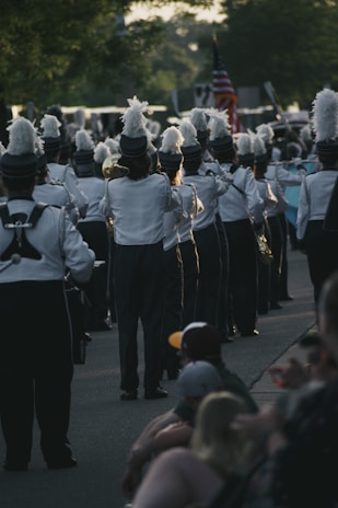 A marching band dressed in uniforms with tall feathered hats is walking down a street. The band members are holding various brass instruments, and an American flag is visible in the background. Onlookers are sitting on the sidewalk, watching the parade.