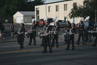 A marching band dressed in uniform, with several members playing drums and cymbals, parades down a street. The background shows a parking lot with parked cars and a two-story building. Several people are sitting or standing near the sidewalk, observing the parade.