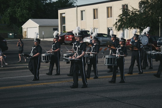 A marching band dressed in uniform, with several members playing drums and cymbals, parades down a street. The background shows a parking lot with parked cars and a two-story building. Several people are sitting or standing near the sidewalk, observing the parade.