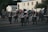 A marching band dressed in uniform, with several members playing drums and cymbals, parades down a street. The background shows a parking lot with parked cars and a two-story building. Several people are sitting or standing near the sidewalk, observing the parade.