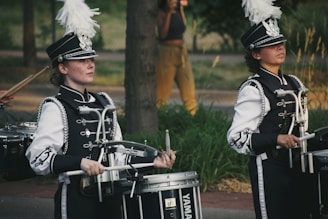 Two marching band drummers wearing intricate black and white uniforms are playing their drums in an outdoor setting. They have decorative hats adorned with large white plumes. The scene suggests a parade or performance.