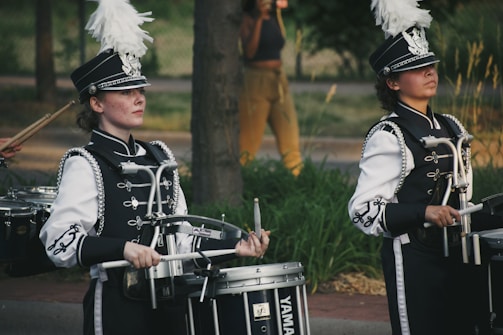 Two marching band drummers wearing intricate black and white uniforms are playing their drums in an outdoor setting. They have decorative hats adorned with large white plumes. The scene suggests a parade or performance.