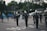 A group of marching band members stand on a street, wearing dark uniforms with white gloves and tall hats adorned with white plumes. One member in the center is dressed in white and holds up a baton. In the background, people carry large blue flags. There are some houses, parked cars, and trees along the street.