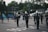 A group of marching band members stand on a street, wearing dark uniforms with white gloves and tall hats adorned with white plumes. One member in the center is dressed in white and holds up a baton. In the background, people carry large blue flags. There are some houses, parked cars, and trees along the street.